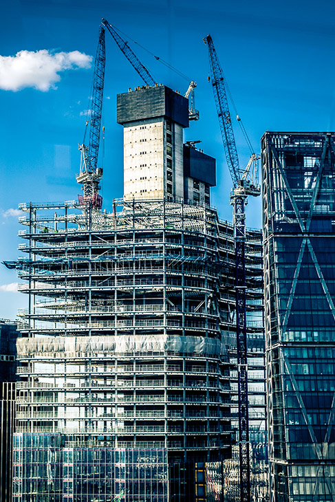 Low angle color image depicting a construction site in London, UK. A housing development is in the process of being created, and multiple cranes are hoisting materials into place. Beyond the cranes and construction equipment is a dazzling sky and cloudscape. Lots of room for copy space.